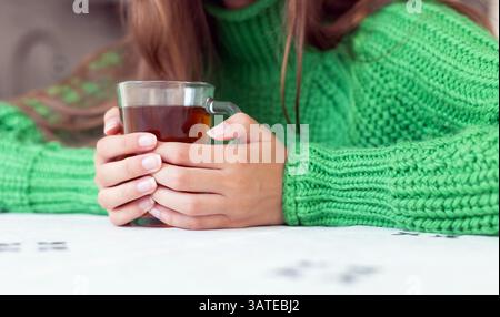 Nahaufnahme der Hände, die ein Glas Tee halten. Nahaufnahme einer Person, die einen grünen Strickpullover trägt und eine klare Glasschale mit Tee hält. Stockfoto