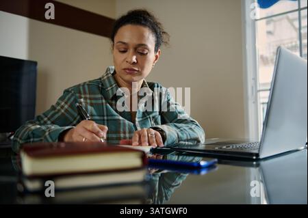 Eine Frau, die an einem Schreibtisch arbeitet und Notizen schreibt, während sie einen Laptop benutzt. Sie arbeitet in einer Home-Office-Umgebung, Multitasking und konzentriert sich auf Produktivität Stockfoto