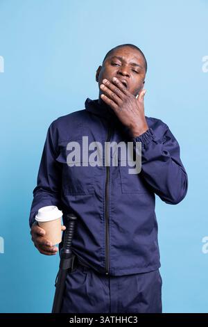 Der afroamerikanische Sicherheitsbeamte fühlt sich nach einer Nachtschicht müde und serviert eine Tasse Koffein, um die Erschöpfung zu bewältigen. Männlicher Offizier, der als Bodyguard arbeitet, gähnt vor blauem Hintergrund. Stockfoto