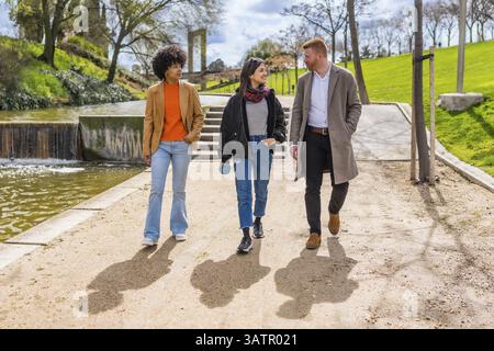Drei junge Berufstätige spazieren gemeinsam in einem sonnigen Park und schlendern entlang eines Pfades an einem Teich Stockfoto