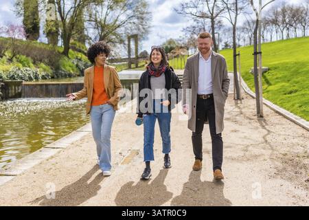 Drei junge Profis machen einen Spaziergang und unterhalten sich während ihrer Mittagspause in einem sonnigen Stadtpark Stockfoto