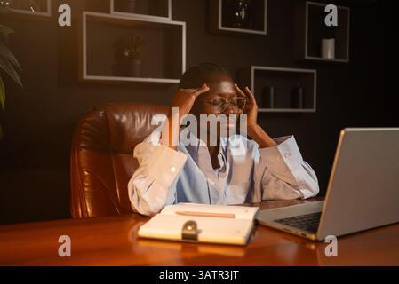Professionelle Geschäftsfrau, die arbeitsbedingten Stress erlebt, Schläfen massiert, während sie bis spät an einem Laptop arbeitet, sitzt am Holzschreibtisch in schwach beleuchteter Büroumgebung Stockfoto
