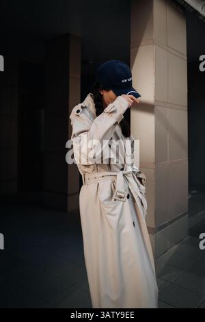 Junge Frau mit trendigem beigem Regenmantel und blauer Mütze, die durch die Stadt geht, moderne Street-Style-Mode Stockfoto