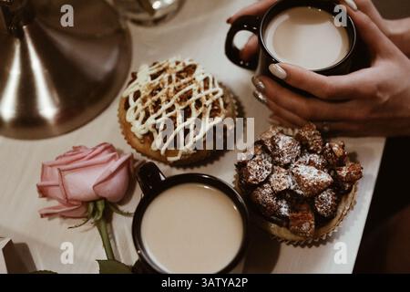 Frauenhände, rosa Rose, zwei schwarze Tassen Kaffee mit Milch und köstliche süße Cupcakes mit Buttercreme und Keksen auf einem Holztisch Stockfoto