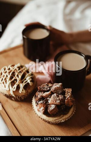 Frauenhände, zwei schwarze Tassen Kaffee mit Milch und köstliche süße Cupcakes mit Buttercreme und Keksen auf einem Holztisch Stockfoto