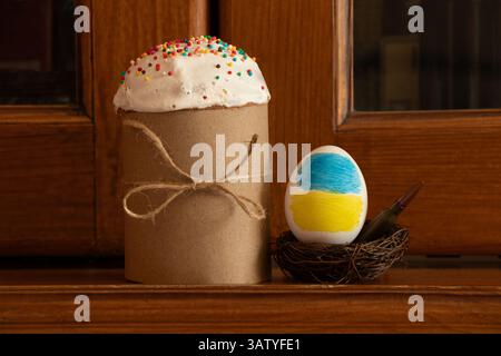 Die Flagge der Ukraine ist auf einem Ei gemalt, das in einem Nest mit einer Kugel liegt, und daneben befindet sich ein Osterkuchen auf einem Holzschrank, Ostern in Ukra Stockfoto