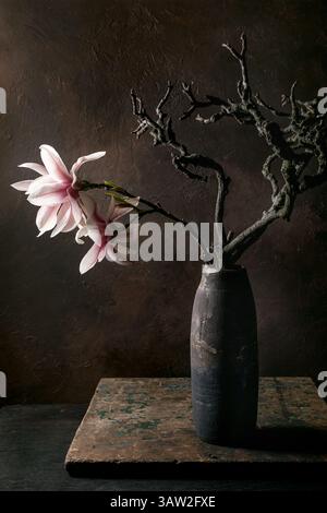 Elegantes Arrangement rosafarbene Magnolienblüten Zweige Ikebana dunkle Keramikvase vor strukturierter brauner Wand. Moody Fine Art Stillleben Komposition Rusti Stockfoto