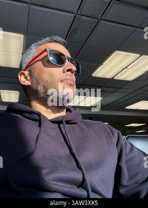 Ein Mann mit Sonnenbrille und schwarzem Hoodie sitzt in einem Raum mit viel Licht Stockfoto