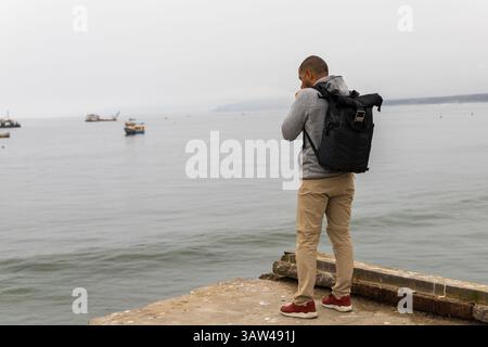 Ein Mann steht auf einem Pier mit Rucksack und Kamera. Das Wasser ist ruhig und es gibt mehrere Boote in der Ferne. Der Mann macht ein Foto von Stockfoto