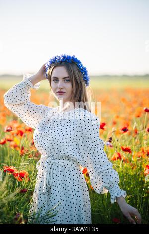 Schöne slawische Frau mit blauer Blumenkrone, die an einem sonnigen Sommertag in einem Mohnfeld posiert Stockfoto