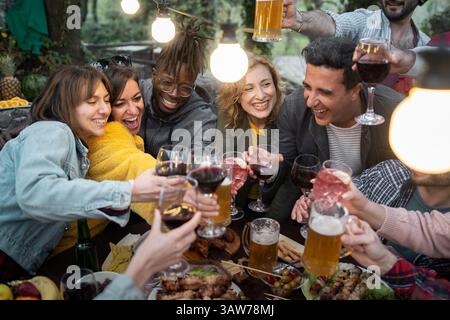Fröhliche, multiethnische Gruppe von Freunden, die während eines Abendessens im Freien mit Getränken locken. Stockfoto