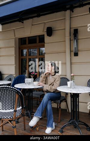 Stilvolle junge Frau genießt Kaffee in einem gemütlichen Café im Freien. Sie war in Jacke und Jeans gekleidet und saß neben dem Marmortisch. Stockfoto