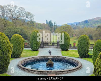Gepflegter symmetrischer Garten mit rundem Wasserbrunnen und getrimmten Hecken. Friedlicher Blick auf das ländliche Anwesen, eingerahmt von Hügeln und Bäumen. Stockfoto
