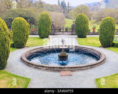 Gepflegter symmetrischer Garten mit rundem Wasserbrunnen und getrimmten Hecken. Friedlicher Blick auf das ländliche Anwesen, eingerahmt von Hügeln und Bäumen. Stockfoto