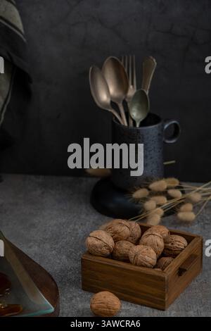 Ein schwach beleuchtetes Stillleben verfügt über eine Holzkiste mit Walnüssen, getrockneten Hasenschwanzgräsern und antikem Besteck in einem dunklen Becher auf einer strukturierten grauen Oberfläche Stockfoto
