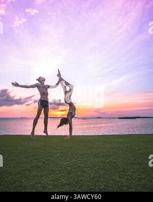 Sportliche Paarsilhouetten für Männer und Frauen, die akrobatische Yoga-Posen auf dem Gras vor dem wunderschönen Sonnenuntergang über dem Meer machen. Der Mann macht Tänzerin-Pose. Stockfoto