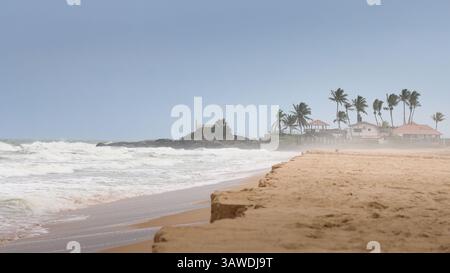 Ruhige Strandszene mit sanften Wellen und hohen Palmen, die ein Küstenresort umrahmen. Der stimmungsvolle Himmel verleiht der ruhigen Umgebung einen Hauch von Drama. Stockfoto