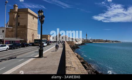 Touristen, die entlang der Cadiz Promenade mit dem Castillo de San Sebastian und der Skyline der Stadt im Hintergrund laufen, Andalusien, Spanien - CADIZ, Spanien Stockfoto