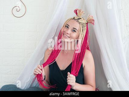 Eine junge Frau lächelt und zeigt ihre einzigartige Frisur mit langen rosa und blonden Zöpfen, die mit kleinen Knoten verziert sind, in einem sanft beleuchteten Raum. Stockfoto