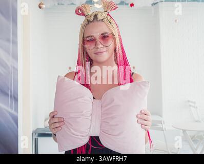 Eine junge Frau posiert mit einem großen, bogenförmigen Kissen und zeigt ihre leuchtend rosa und blonden Zöpfe. Stockfoto