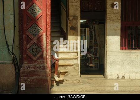 Havanna, Kuba 2024 April 17 - Ein Mann sitzt auf der Treppe im Schatten vor einem Barbershop in der Nachbarschaft, während ein anderer sich innen einen Haarschnitt gönnt. Straßenszene o Stockfoto