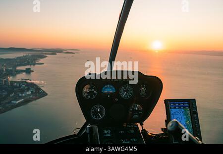 Hubschrauberrundblick über das Cockpit bei Sonnenuntergang über die Küstenstadt mit ruhigem Wasser und fernen Inseln Stockfoto