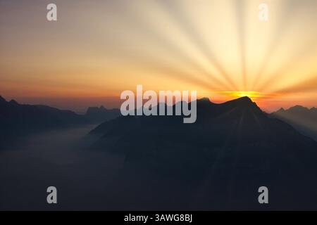 Sonnenstrahlen über Schweizer Berggipfeln bei Sonnenuntergang mit Tal und See, buntes Landschaftsbild Stockfoto