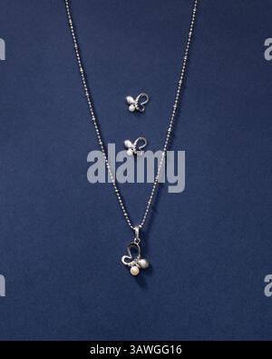 Silberne Schmetterlings-Halskette und Ohrring mit weißen und grauen Perlen, isoliert auf einem dunkelblauen strukturierten Hintergrund. Minimalistischer Produktfoto Stockfoto