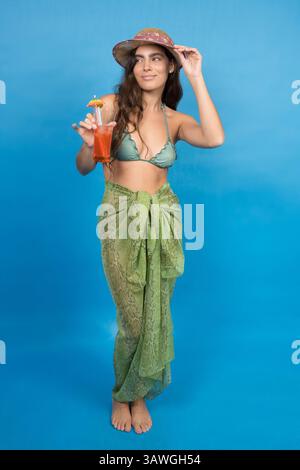 Studio-Aufnahme einer attraktiven jungen Frau, die Sarong, Bikinioberteil und Strohhut trägt und tropischen Cocktail vor blauem Hintergrund hält Stockfoto