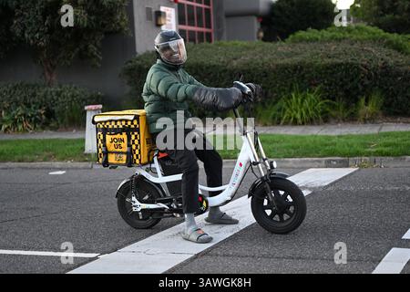 Fröhlicher Essenskurier, der an einer Kreuzung auf einem weißen E-Bike sitzt, mit gelbem Aufbewahrungsbehälter für hungrige Panda auf der Rückseite des Fahrrads Stockfoto