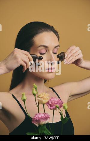 Frau, die Hautpflegeroutine mit Gesichtsrollen und Blumen praktiziert Stockfoto