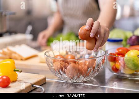 Nahaufnahme einer Person, die frische braune Eier aus einer Glasschale in einer kommerziellen Küche auswählt. Zu den umliegenden Zutaten gehören Gemüse, Brot und Obst Stockfoto