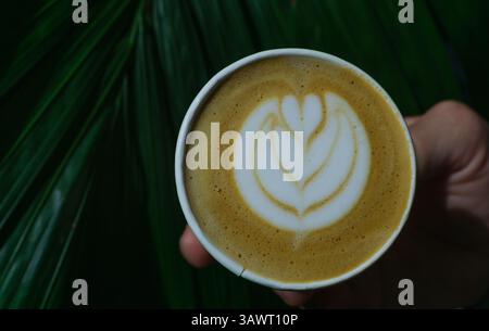 Bei einer Nahaufnahme wurde ein Papierbecher zum Mitnehmen mit einem Latte gedreht, der zarte herzförmige Latte Art im cremigen Schaum zeigt. Ein Hinweis auf eine Hand hält t Stockfoto