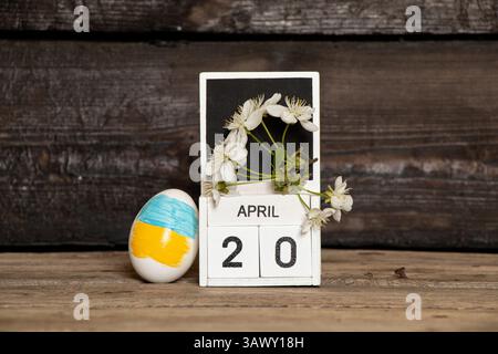 Osterei mit ukrainischer Flagge neben Kalender mit Datum vom 20. April und Zweig mit Kirschblüten auf hölzernem Hintergrund Stockfoto