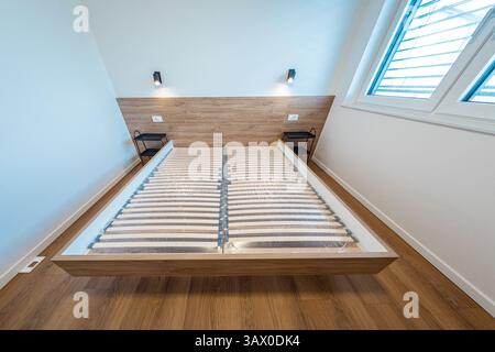 Ein modernes Schlafzimmer mit Holzrahmen, Lattenrost und ohne Matratze, hat Hartholzböden, weiße Wände, ein Fenster mit Jalousien, ein Schreibtisch, ein Rot Stockfoto