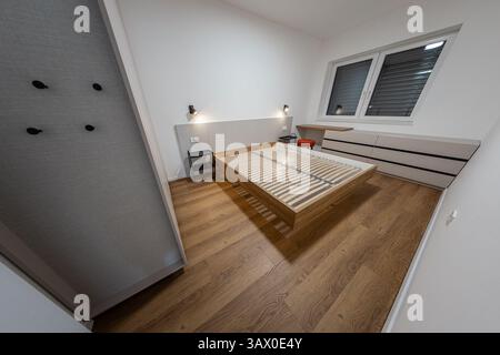 Ein modernes Schlafzimmer mit Holzrahmen, Lattenrost und ohne Matratze, hat Hartholzböden, weiße Wände, ein Fenster mit Jalousien, ein Schreibtisch, ein Rot Stockfoto