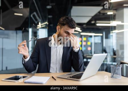 Ein Geschäftsmann in einem Anzug, der Kopfschmerzen hat, zieht seine Brille an seinem Schreibtisch ab. Er benutzt einen Laptop, arbeitet wahrscheinlich hart und fühlt sich gestresst. Stockfoto