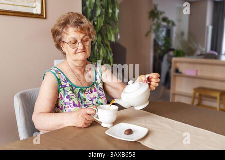 Seniorin mit kurzen Haaren und Gläsern, die allein zu Hause sitzen, Tee mit süßen Süßigkeiten auf dem Tisch trinken, ungesunde Essgewohnheiten und Risiken des Zuckerkonsums Stockfoto