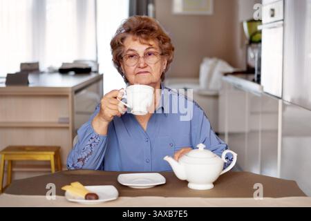 Ältere Frau in blauer Kleidung sitzt allein zu Hause, trinkt Tee und trinkt trauriges Frühstück, sieht nachdenklich und emotional aus in ruhiger Morgenstunde Stockfoto