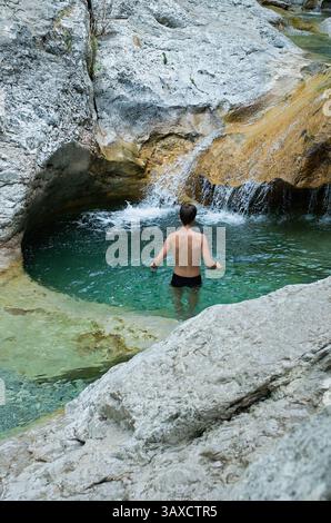 Ein junger Mann schwimmt in einem türkisfarbenen Natursteinpool, umgeben von zerklüfteten Klippen, mit einem kleinen Wasserfall, der ins Wasser stürzt. Perfekt für Themen von Stockfoto