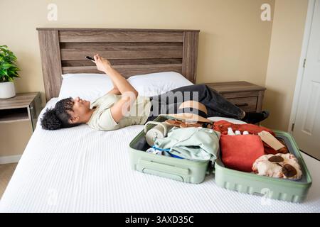 Die Frau liegt auf dem Bett und lächelt am Telefon neben dem gepackten Koffer Stockfoto