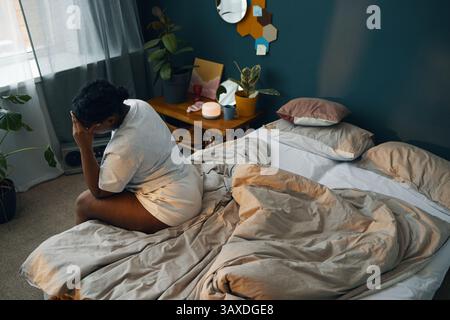 Schwarze Frau, die auf ungepflegtem Bett sitzt und den Kopf in einer nachdenklichen Pose hält. Das Schlafzimmer verfügt über Pflanzen, Wanddekorationen und eine sanfte Atmosphäre, die von einem Wind in der Nähe beleuchtet wird Stockfoto