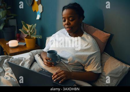 Frau, die sich im Bett zurücklehnt und ein Getränk in der Hand hält und am Laptop arbeitet, komfortabel umgeben von Pflanzen und Dekor in einem gemütlichen Zimmer Stockfoto
