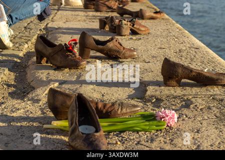 Nahaufnahme und weite Aussicht auf Shoes am Donauufer-Denkmal in Budapest mit Bronzeschuhen, Blumen und Kerzen entlang der Uferpromenade Stockfoto