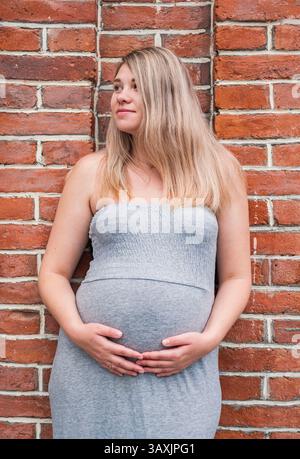 Die junge schwangere Frau steht selbstbewusst und hält sich an eine Ziegelwand. Stockfoto