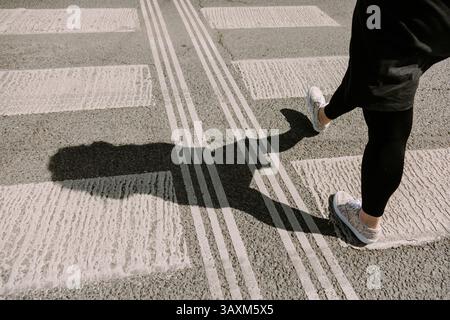 Frau, die über einen Fußgängerübergang geht, mit einem klaren Schatten auf dem Gehweg. Perfekt für Themen wie Stadtleben, Unabhängigkeit, Bewegung und Stockfoto