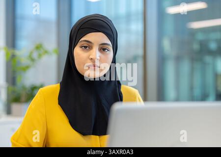 Eine Frau mit schwarzem Hijab und gelber Bluse posiert mit ernstem Ausdruck in einem Büro. Der Laptop deutet an, dass sie arbeitet. Stockfoto