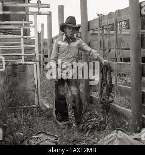 Monochromes Vintage-Porträt eines Rodeo-Cowboys mit Lederbeinen und Lehnen an einer Rutsche in einer Schrift über Stone, Alberta, Kanada. Um 1983 Stockfoto