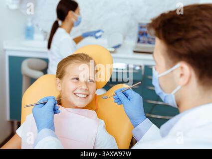 Männlicher Arzt im weißen Mantel mit Gummihandschuhen und Schutzkleidung Maske behandelt Zähne von lächelnden Mädchen im Stuhl Stockfoto