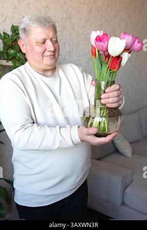 Lächelnder älterer Mann in einem hellen Pullover, der eine klare Glasvase mit roten, weißen und rosa Tulpen in einem gemütlichen Wohnzimmer hält, umgeben von Natur Stockfoto